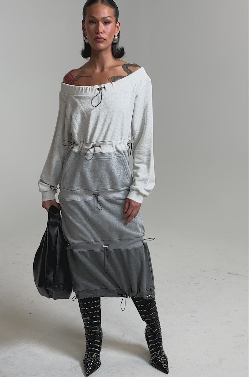 A video of a woman in the TIED UP RIGHT NOW MAXI DRESS, an off-the-shoulder grey colorblock sweatshirt dress with drawstring details, paired with studded knee-high boots, bold earrings, and a black bag against a plain background.