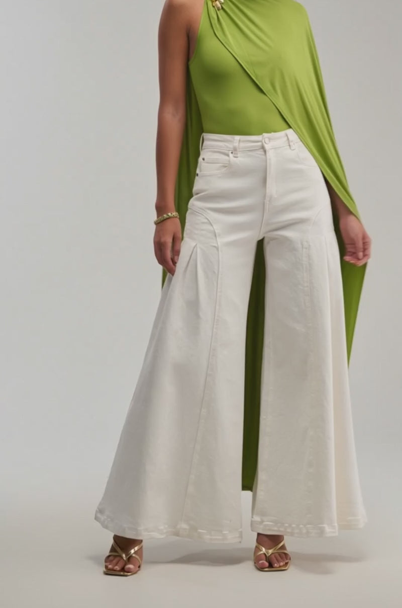 A video of a woman in the NOT YOURS CAPE BODYSUIT IN CHARTREUSE with draped detail, paired with high-waisted wide-leg white pants and gold sandals against a plain light backdrop.