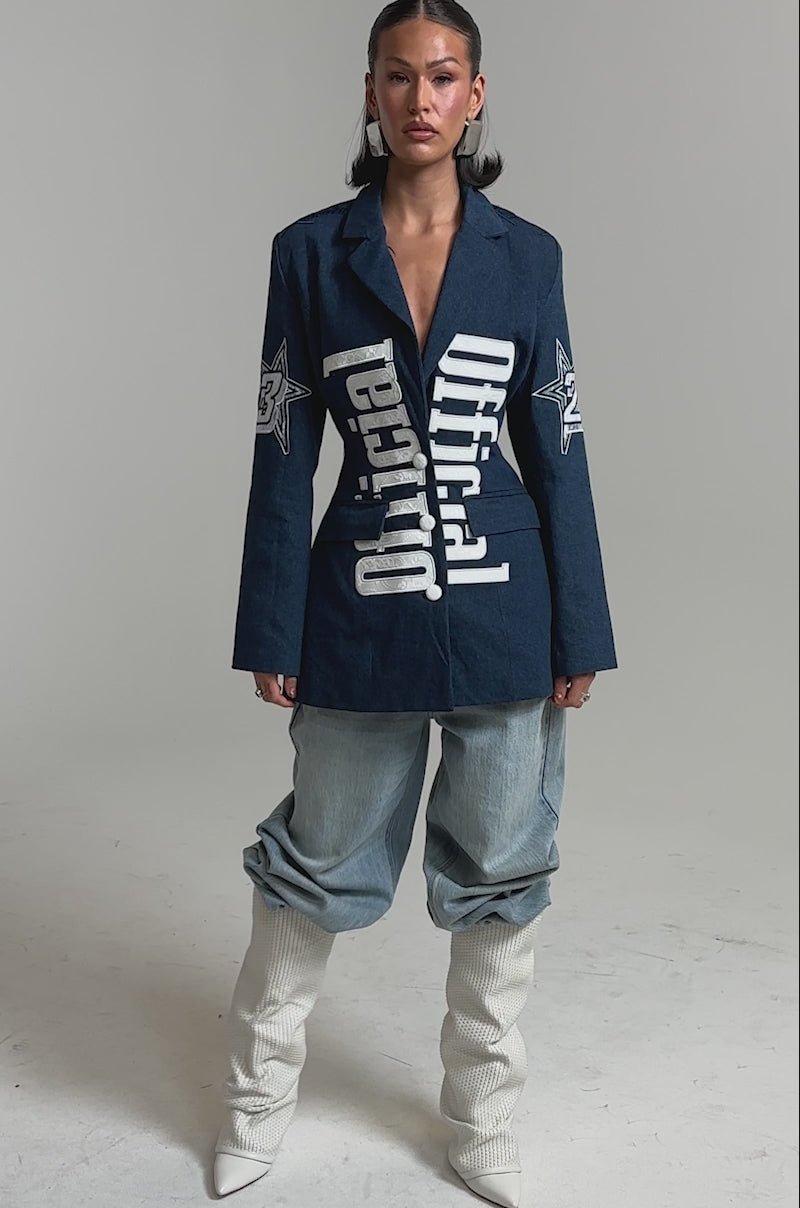 A video of a woman in the ONE OF ONE BACKLESS DENIM BLAZER featuring bold "Official" lettering, styled with light blue baggy jeans and white slouchy boots.
