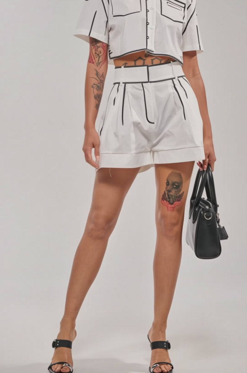 Video of woman modeling the BLANK SPACE SHORT—high-waisted festival shorts with black trim—and a matching cropped top, displaying tattoos on her arms and thigh, while holding a black handbag at her side.