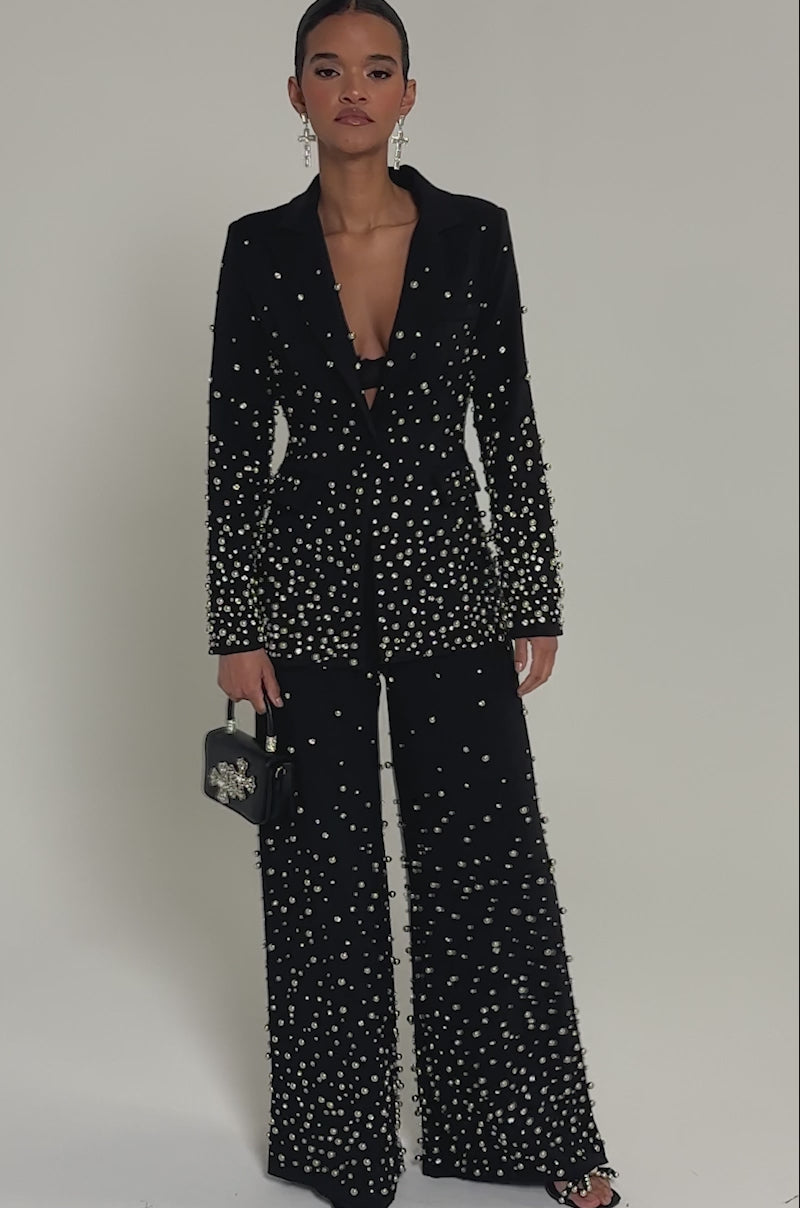 Video of woman modeling the GET CLOSER EMBELLISHED BLAZER with matching pearl-adorned pants and a jeweled black bag. Her slicked-back hair, bold cross earrings, and confident expression complete the look.