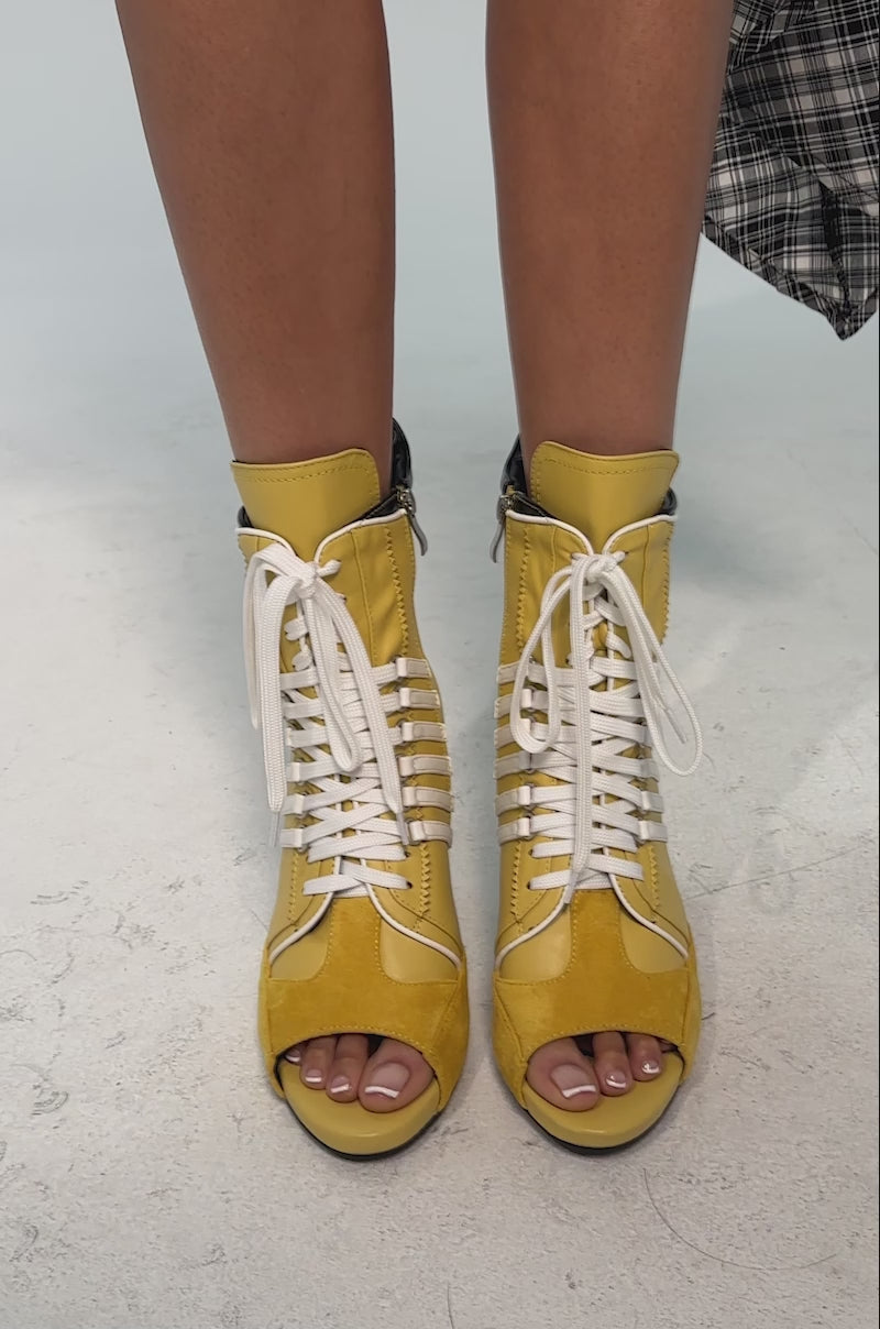 A video of a model wearing the AZALEA WANG EOWYN Mustard Yellow Open Toe Stiletto Bootie featuring white laces, horizontal white side straps, a black back detail, and a high-heeled ankle boot design.