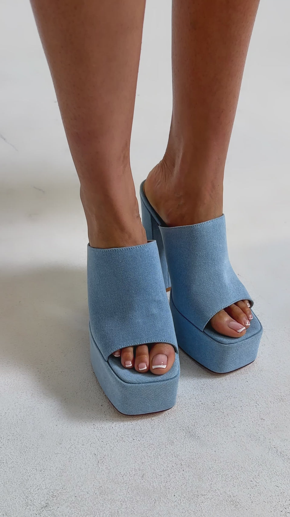 Video of woman modeling the AKIRA GOLD FIKILE DENIM CHUNKY SANDAL, featuring light blue denim, open square toes, and chunky block heels, stands against a neutral background.