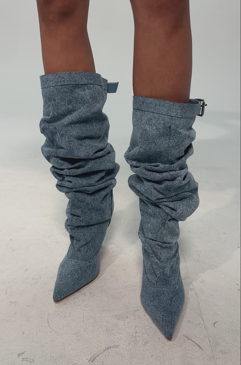 A video of a model wearing the AZALEA WANG DARIA DENIM STILETTO BOOT. It is a single high-heeled, pointed-toe slouchy light blue denim boot featuring a decorative strap and buckle near the top.