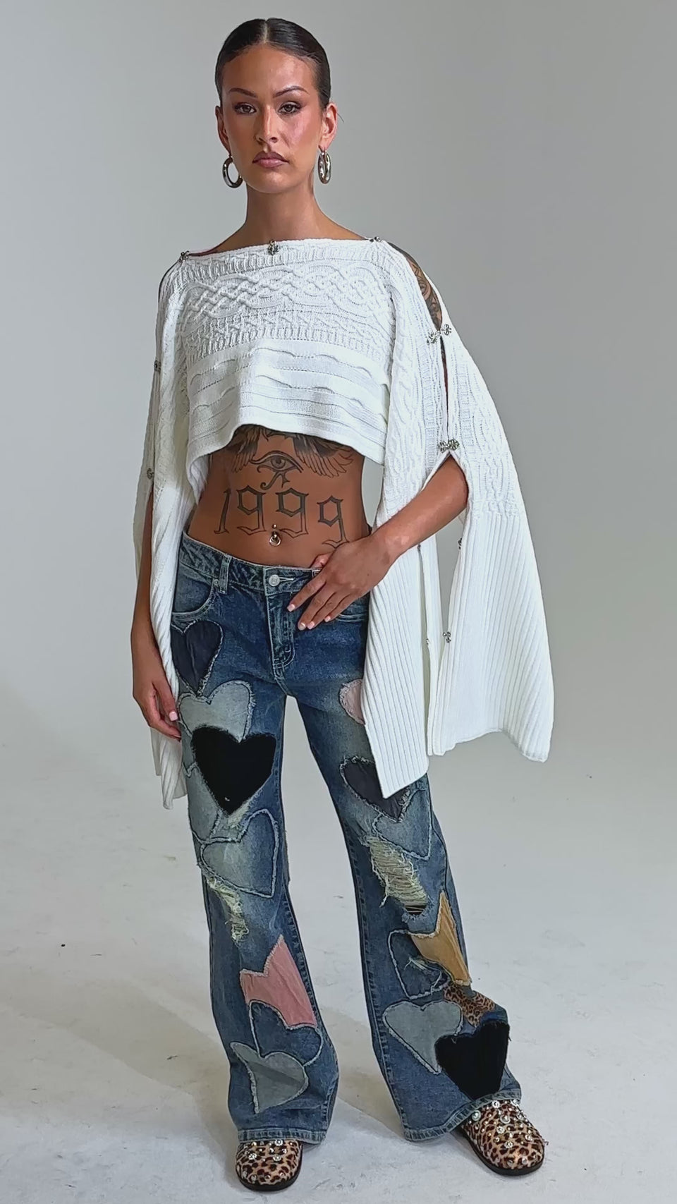 Video of woman modeling the LOVE ME HARD RELAXED DENIM with large, colorful heart patches, this person styles the bold jeans with leopard-print shoes, a white open cardigan, and shows off a midriff tattoo.