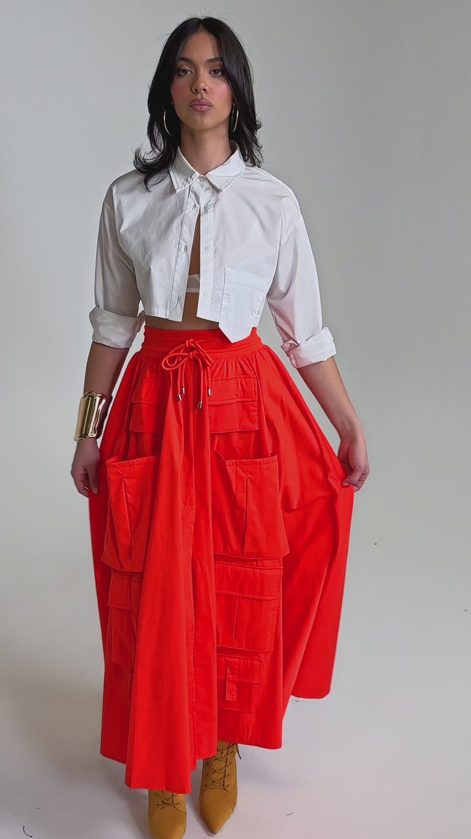 A video of a woman wearing a cropped white button-up shirt, mustard yellow heeled boots, and the AMELIA CARGO POCKET MAXI SKIRT IN RED against a plain background.