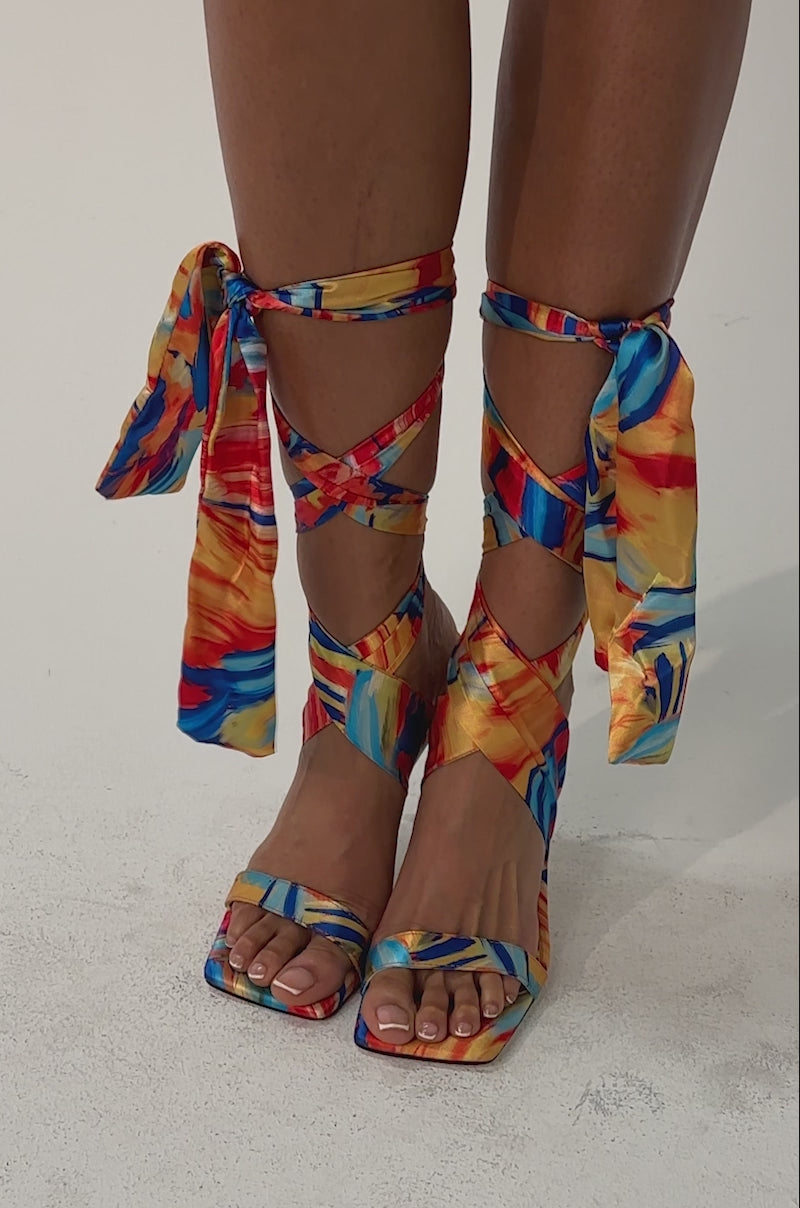 Video of woman modeling the AZALEA WANG JOHA ORANGE MULTI PRINTED SATIN WRAP UP STILETTO—high-heeled sandals with open square toes and long, multicolored wraparound satin straps—against a plain light background.