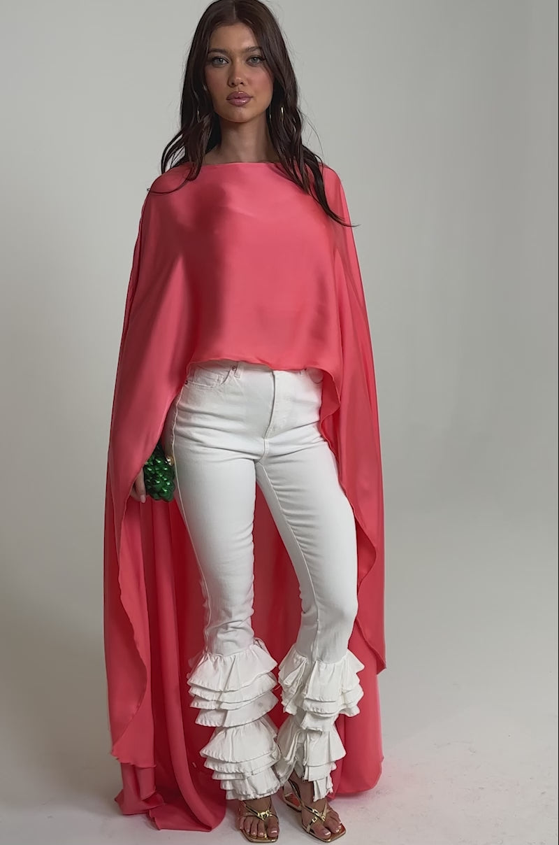 Video of woman modeling the SO CHIC CAPE BLOUSE, paired with white ruffled pants and gold heels, holding a small green textured clutch against a plain, light background.