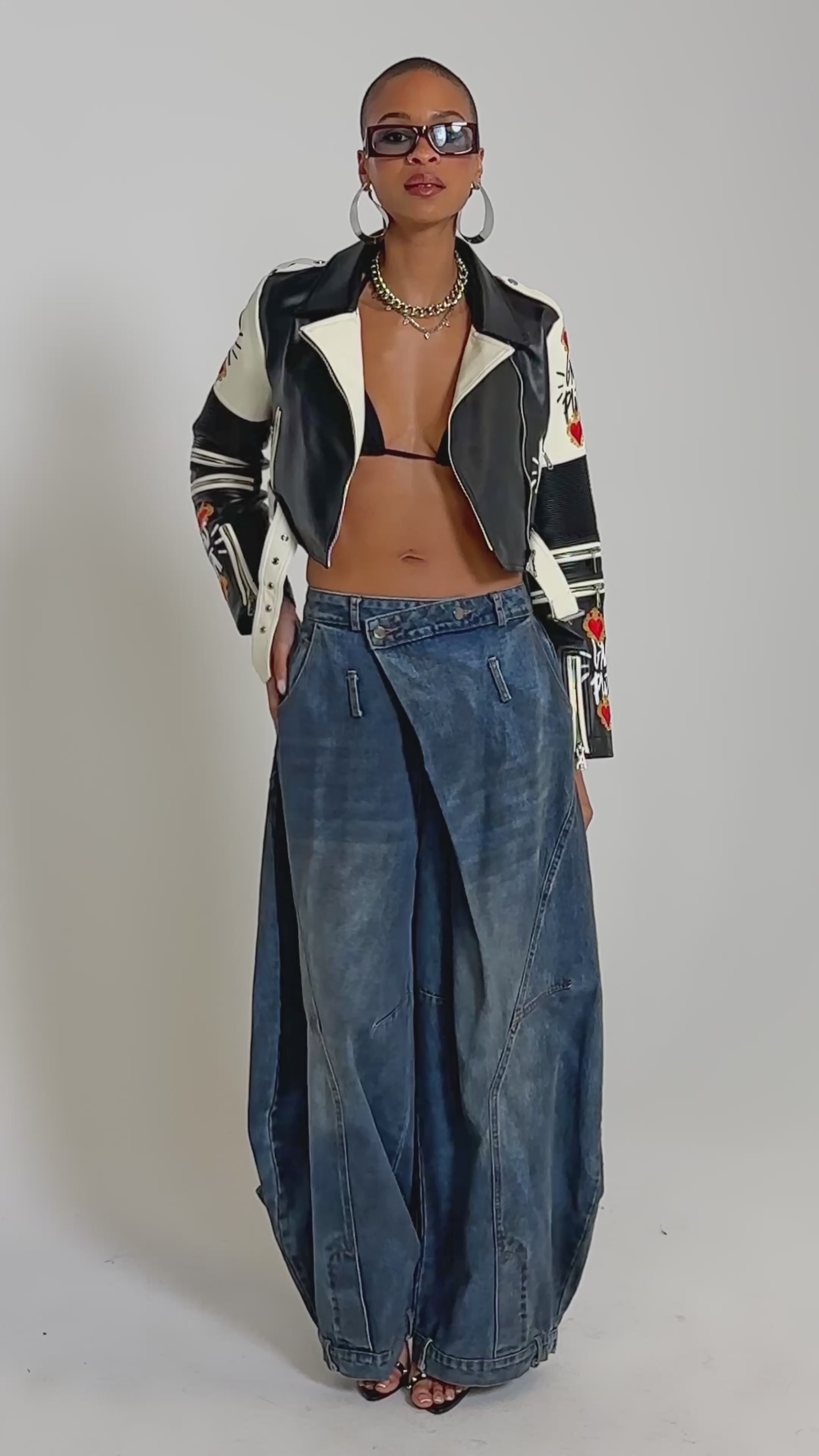 Video of woman modeling the SUNNY GRAFFITI MOTO cropped biker jacket with graphic designs, paired with blue jeans, hoop earrings, layered necklaces, sunglasses, and carries a zebra-print clutch.
