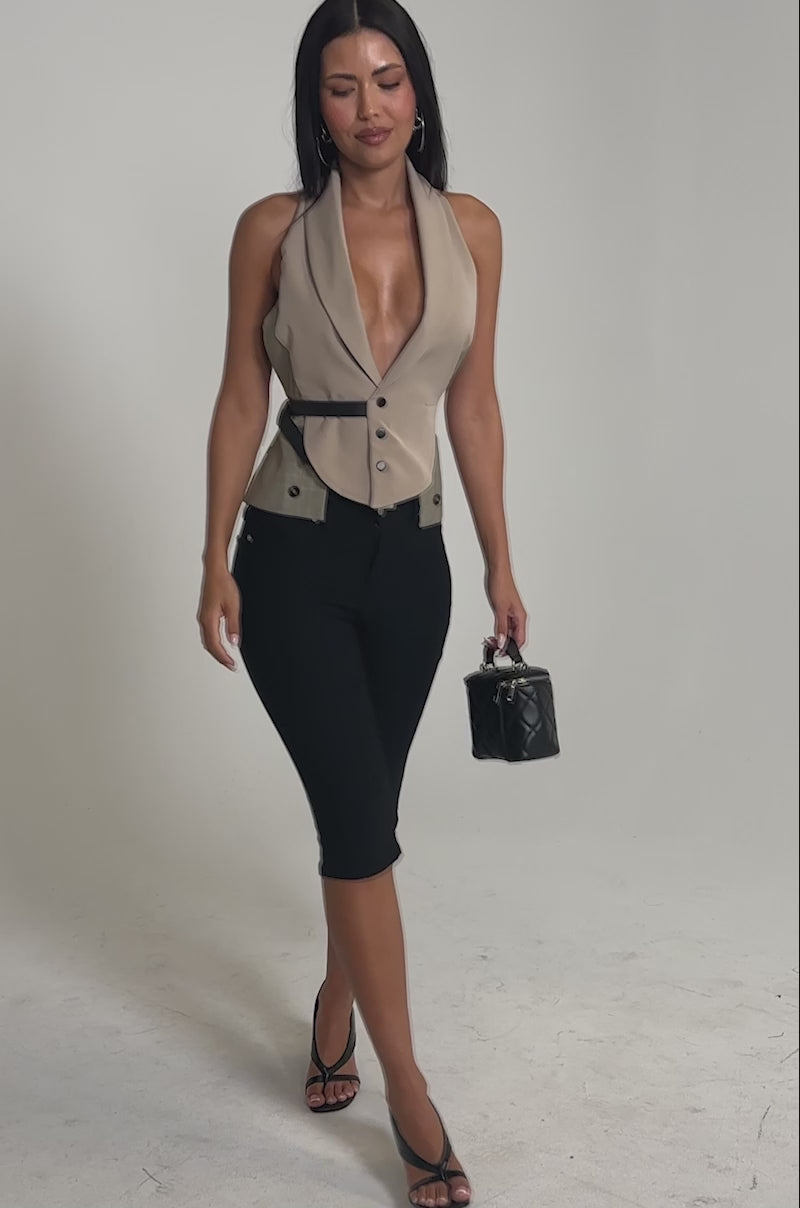 Video of woman modeling the DENIM MUSE CAPRI, pairing these black high-rise capri pants with a taupe sleeveless belted vest, black high-heeled sandals, and a small black handbag.