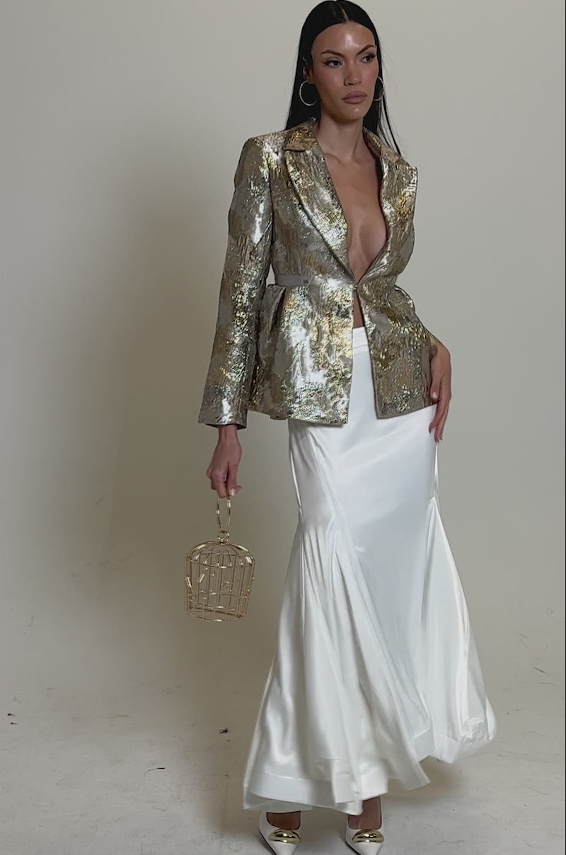 A video of a woman in the CANARY PEPLUM BLAZER IN CHAMPAGNE over a flowing white skirt, holding a gold cage purse and wearing pointed heels. Her long, straight dark hair completes the chic look against a plain background.