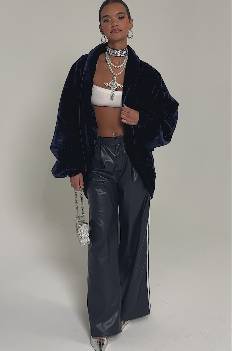 Video of woman modeling the LOLA COCOON COAT IN VELVET (royal blue), navy leather pants, a white crop top, layered pearl necklaces with a jeweled cross, hoop earrings, and carrying a metallic silver handbag.