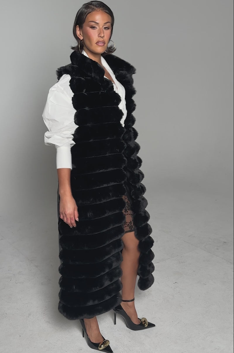 A video of a woman posing confidently against a light background in the POPPY FAUX FUR VEST layered over a white blouse and black lace skirt, accessorized with gold hoop earrings, a wide belt, and black heels.