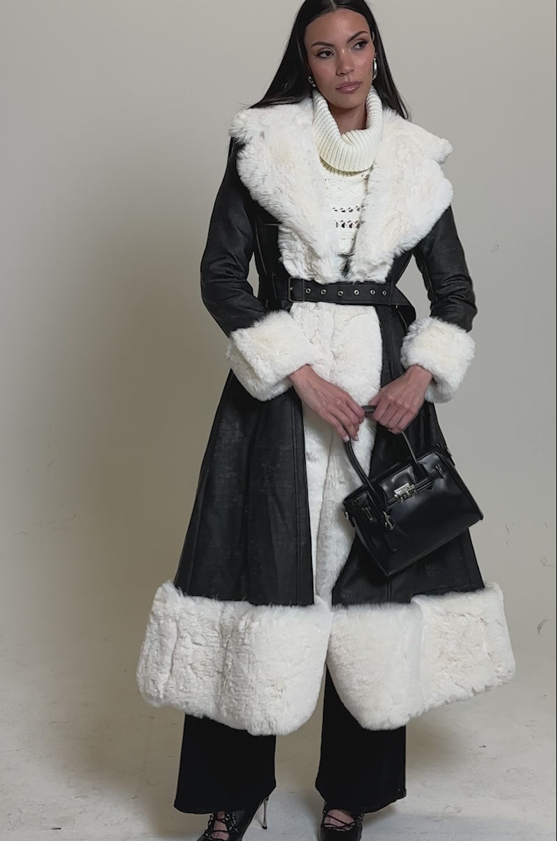 A video of a woman with long straight hair and a confident pose wearing the VIVIAN FUR TRIM TRENCH IN GREY, paired with a cream turtleneck sweater, dark pants, black heels, and holds a black handbag.