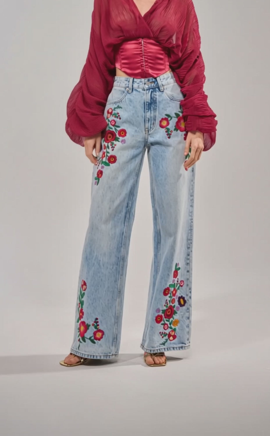 Video of woman modeling the GIVE ME FLOWERS WIDE LEG DENIM, featuring colorful floral embroidery on the thighs and lower legs, styled with a sheer red wrap top over a satin blouse. She has long blonde hair and finishes the look with sandals.