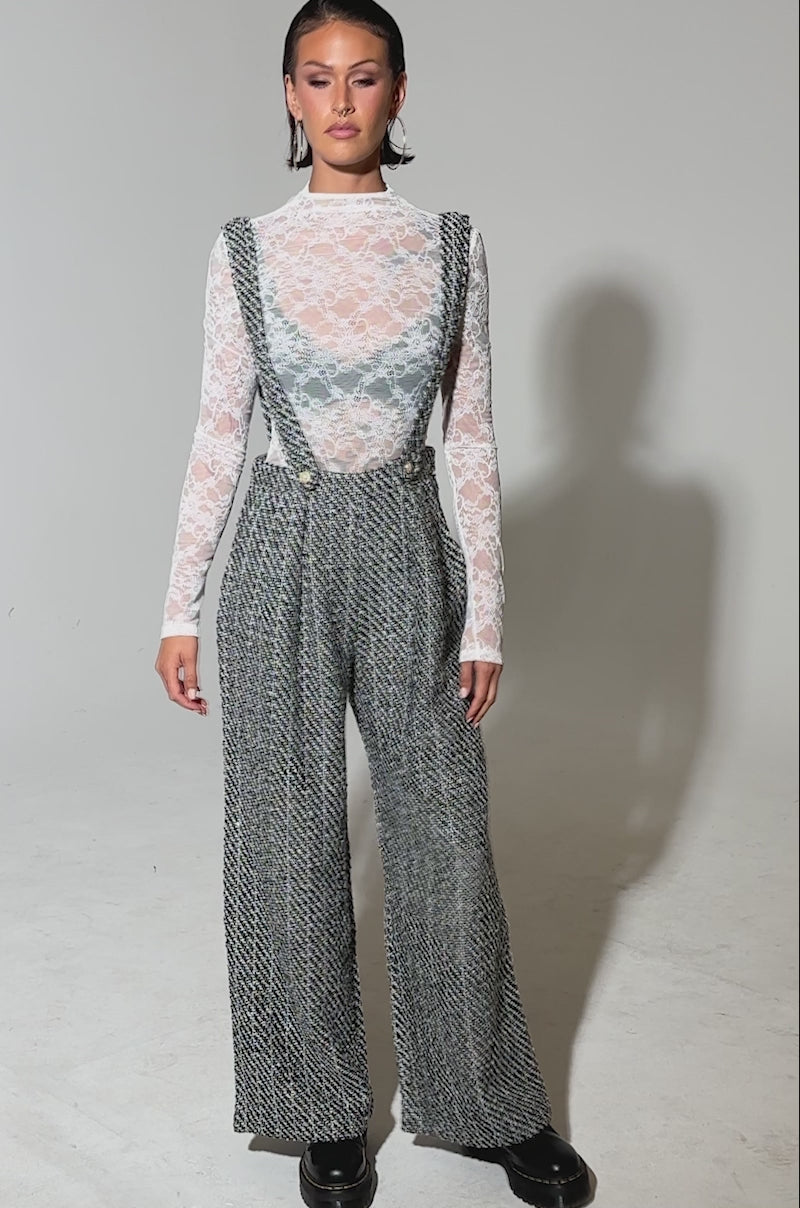 A video of a woman wearing the ALESSANDRA TWEED OVERALLS, featuring pearl button straps over a sheer white lace long-sleeve top, paired with black chunky platform shoes.