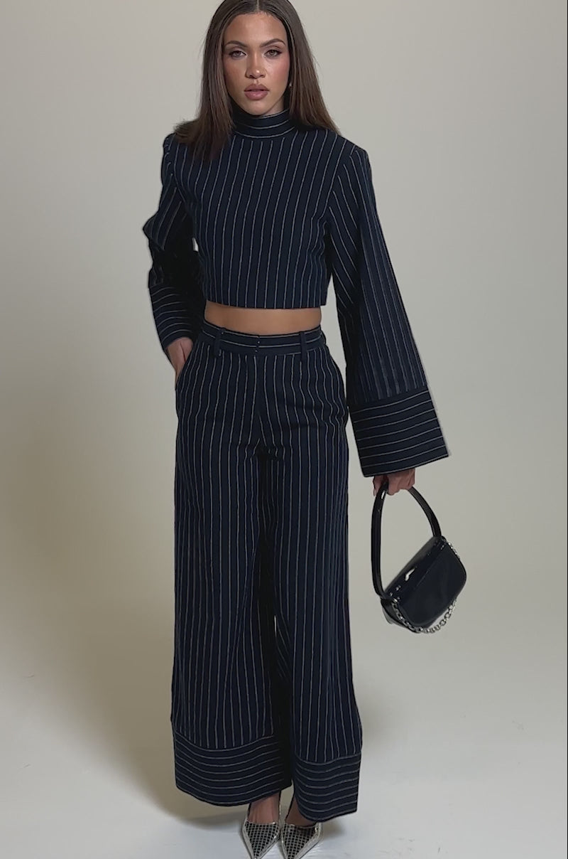 Video of woman modeling the STRICTLY BUSINESS PINSTRIPE TROUSER set—navy blue, wide-leg pants and a cropped long-sleeve top. She accessorizes with a black handbag and silver pointed heels, posing against a plain light background.