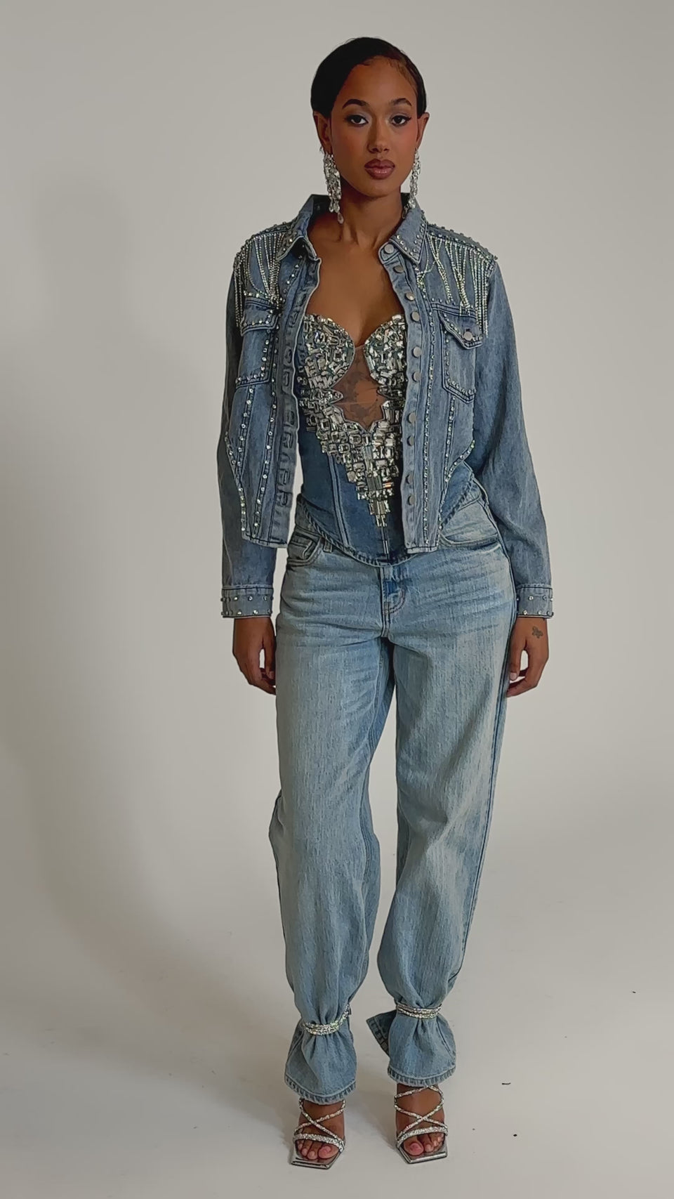A video of a woman wearing the HOOKED ON YOU EMBELLISHED DENIM JACKET over a sheer, sparkly top, paired with light blue high-waisted cuffed jeans, silver heels, and large earrings.