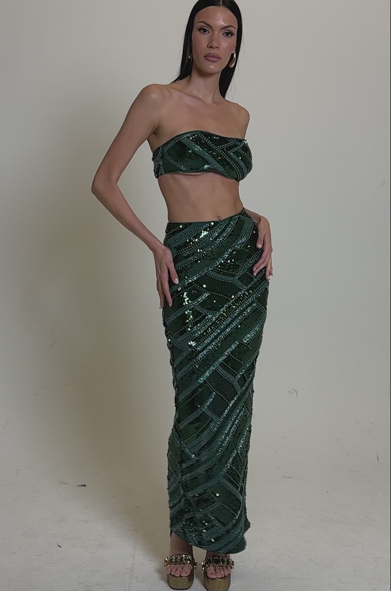 A video of a woman in the HOTTEST GIRL IN THE CLUB SEQUIN BANDEAU, a strapless crop top, paired with a matching high-waisted, floor-length geometric skirt. She wears gold platform heels and has long, straight dark hair.