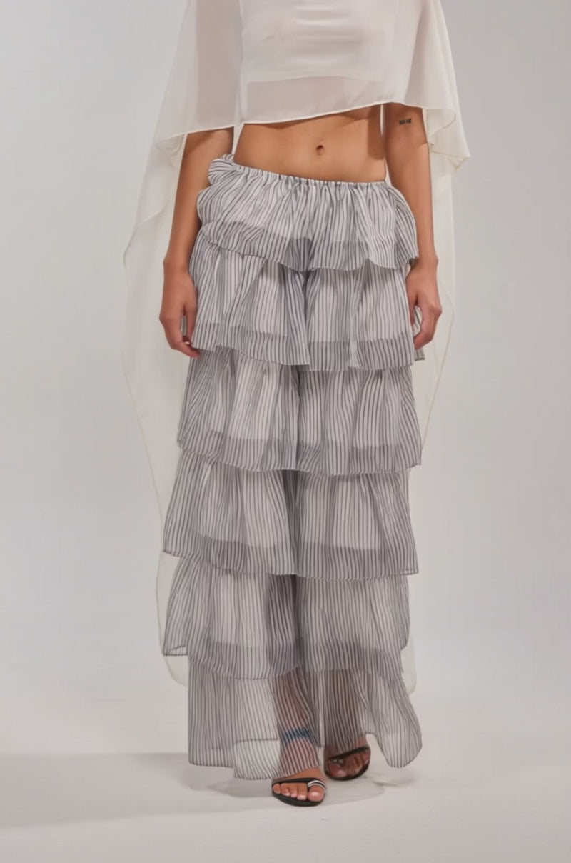 Video of woman modeling the IT GIRL BLOUSE IN IVORY, a sheer chiffon cape top with a high-low drape, paired with a ruffled striped pant, chunky silver necklace, and open-toe heels against a plain backdrop—perfect for festivals.