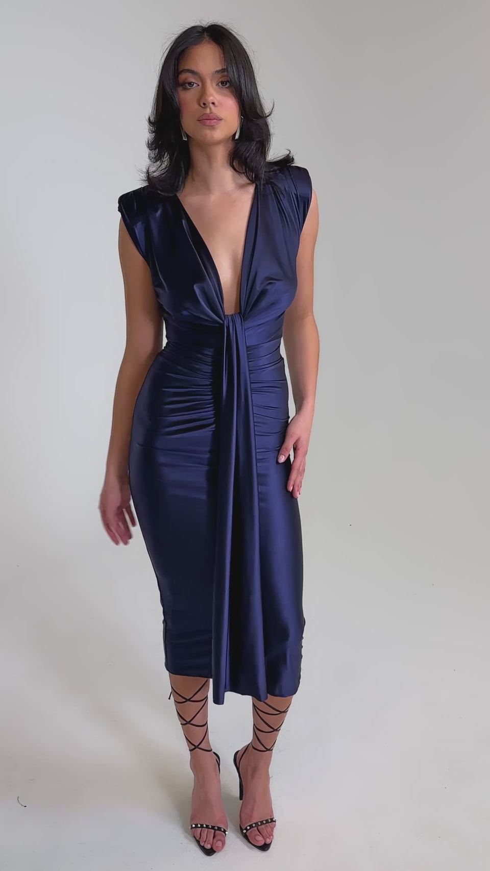 A video of a woman wearing the FLY ME OUT SATIN MAXI DRESS, a sleeveless deep v-neck purple satin maxi with ruched detailing and knot-front draped tie, styled with strappy black heels.