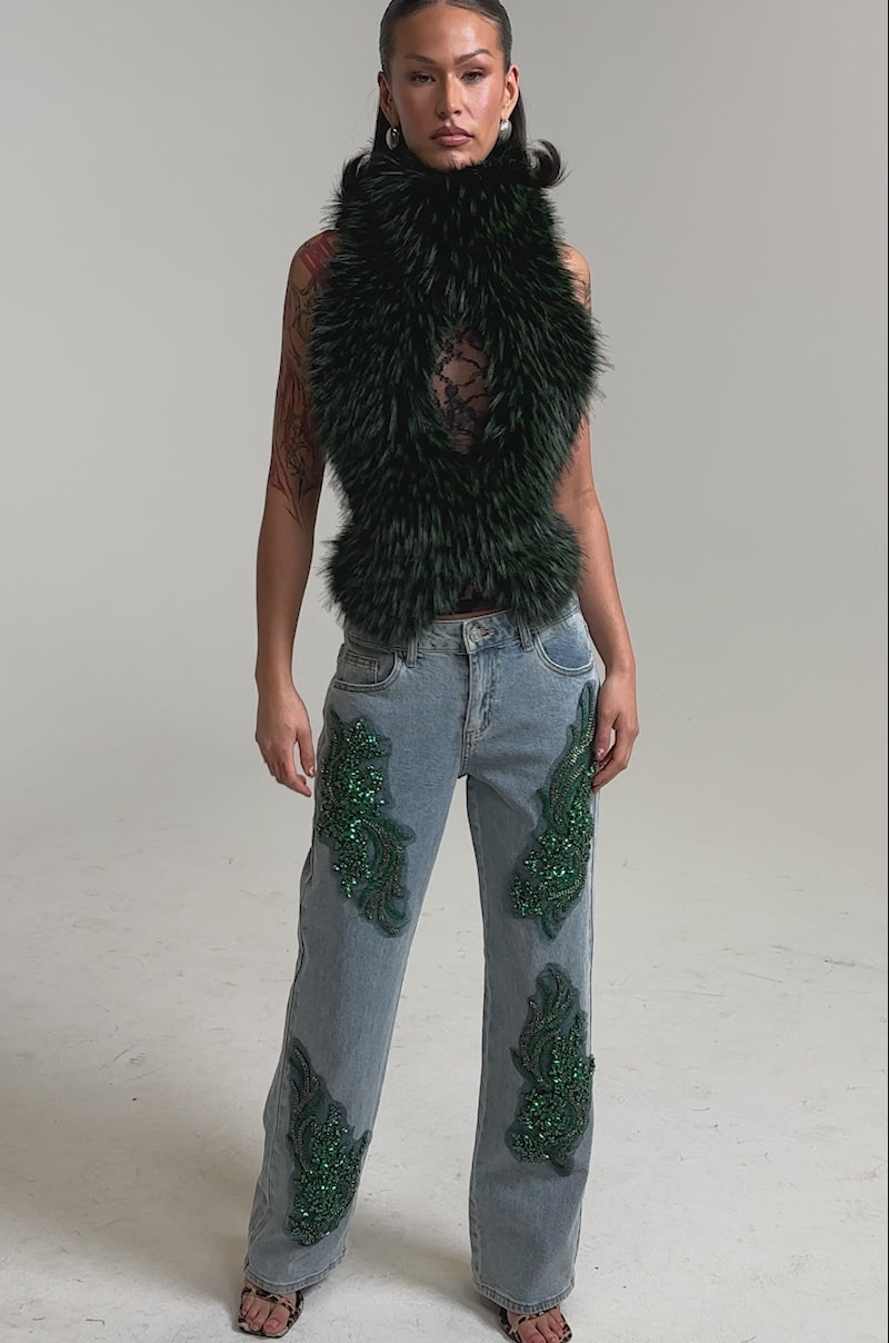 A video of a woman in a black faux fur vest, sheer lace top, and the high-rise BILLIONAIRE BELLE RHINESTONE JEANS embellished with light-wash denim and green sequins.