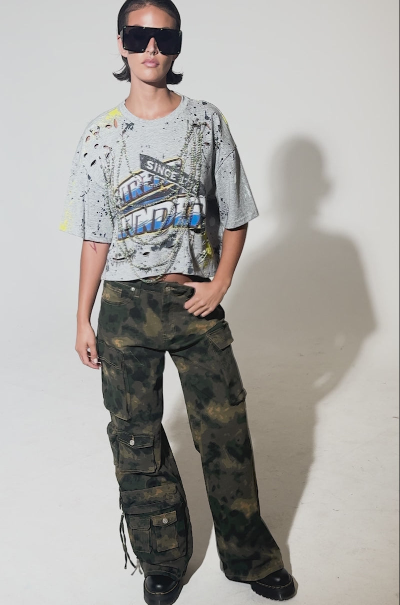 A video of a person wearing large sunglasses, a distressed gray graphic tee, and the WASHED UP CARGO CAMO PANT—loose-fitting with a wide-leg silhouette—against a plain light background.