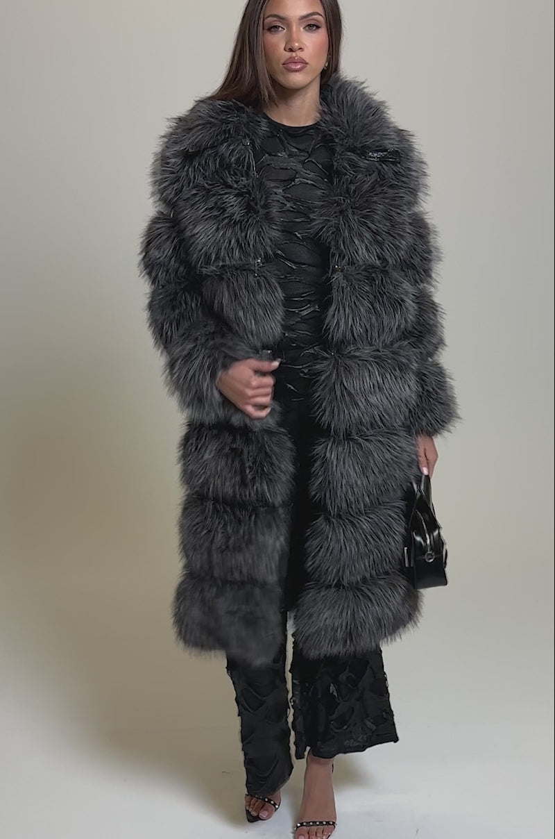 Video of woman modeling the NOELLE ZIP OFF TRENCH, a long plush gray faux fur coat, over a black textured dress with heeled sandals and a small black handbag. 