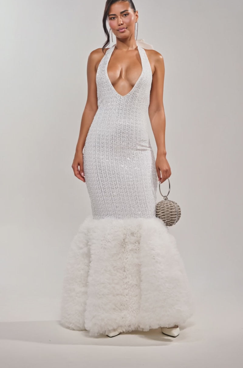 Video of woman modeling the SHOWTIME SEQUIN MAXI DRESS, a halter mermaid gown with a deep V-neckline and ruffled skirt. She pairs it with a round silver handbag, white pointed shoes, and a sleek ponytail for an elegant formal look