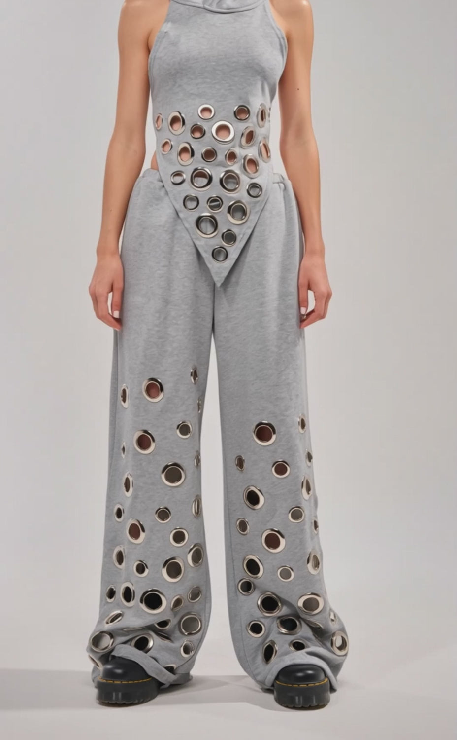 Video of woman modeling the CHECK IT OUT GROMMET JOGGER—high-rise pants with oversized grommets and large circular cutouts—paired with a sleeveless gray hooded top. She completes the look with sunglasses and chunky black shoes