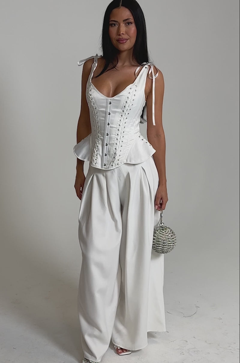 A video of a woman in the FEELING THE VIBES CORSET and wide-leg white trousers, holding a round silver handbag. 
