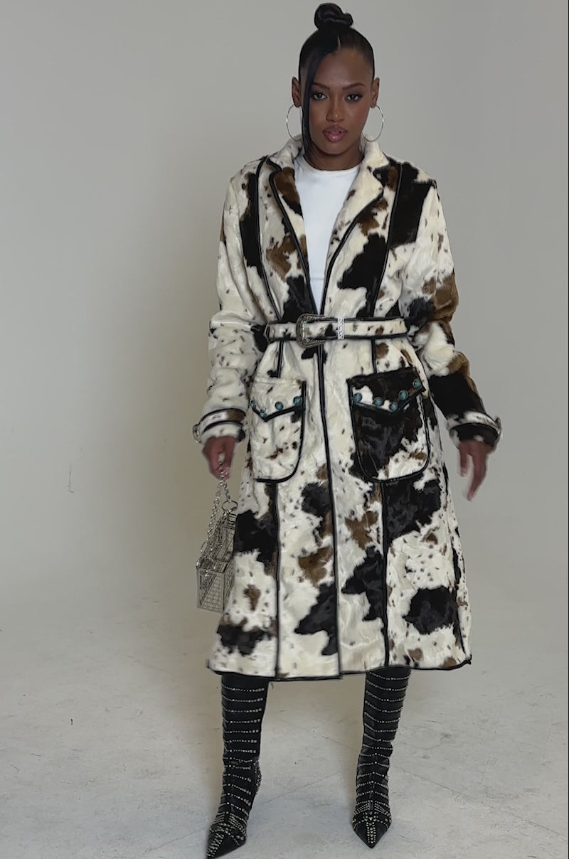 Video of woman modeling the RIDE EM COW PRINT TRENCH faux fur coat, paired with black studded knee-high boots, hoop earrings, a small metallic handbag, and a high bun hairstyle.