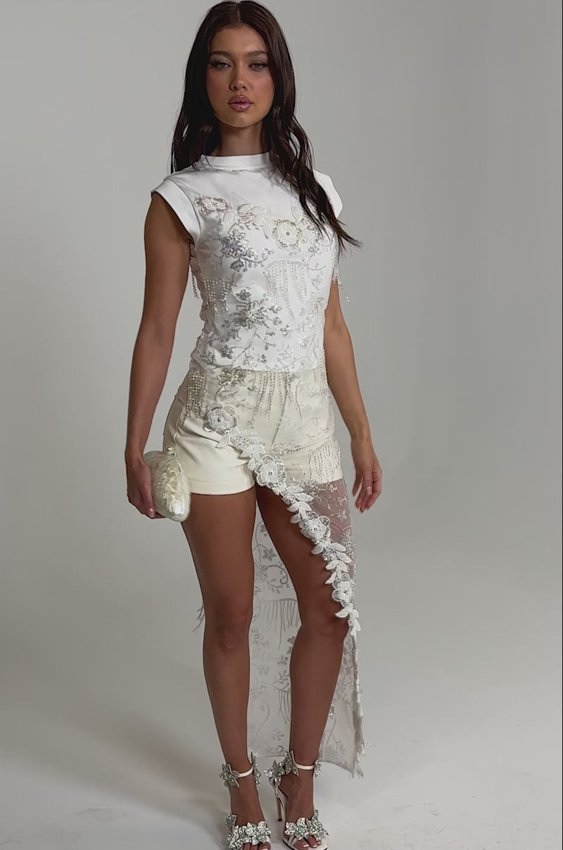 Video of woman modeling the SHINE BRIGHT BLOUSE, a sheer white asymmetrical top with floral embroidery, paired with cream shorts, white floral heels, and a small white clutch against a plain light-colored background.