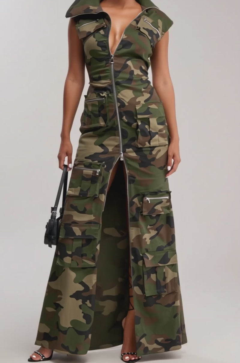 Video of woman modeling the NOW YOU SEE ME CAMO PRINT MAXI DRESS featuring a front zipper, thigh-high slit, and cargo pockets. She completes the look with strappy heels, sunglasses, and a small black handbag