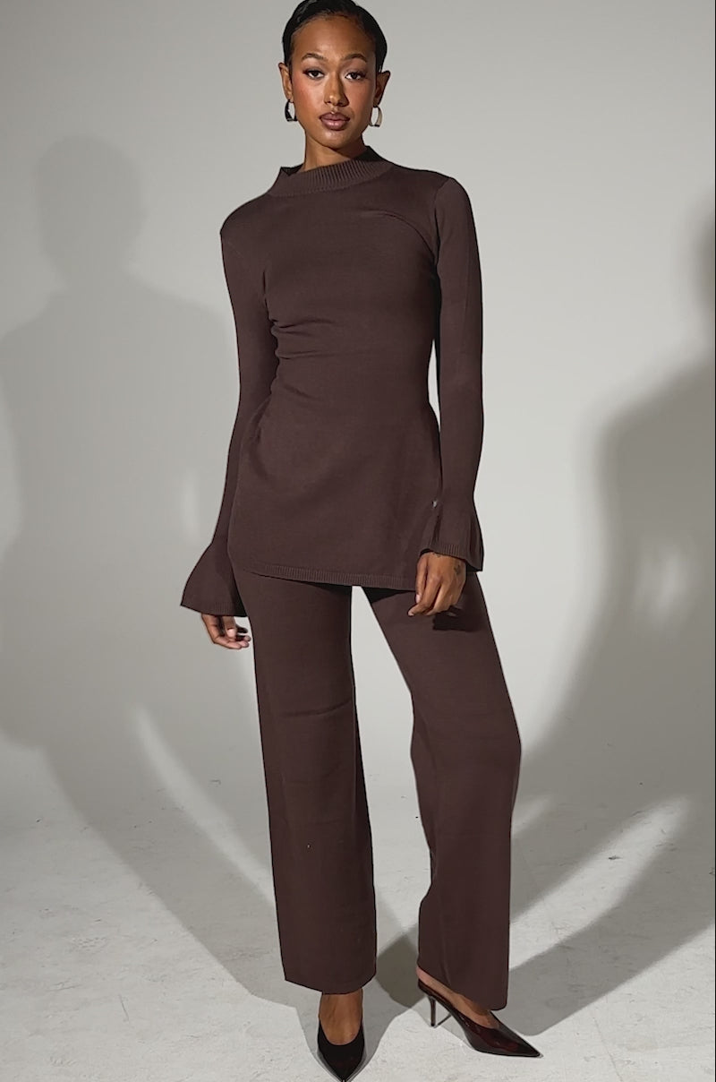 A video of a woman posing confidently against a light background in the GET COZY WITH ME WIDE LEG PANT and a matching brown knit sweater, paired with black heels and short hair. The outfit is crafted from stretchy knit fabric for comfort and style.