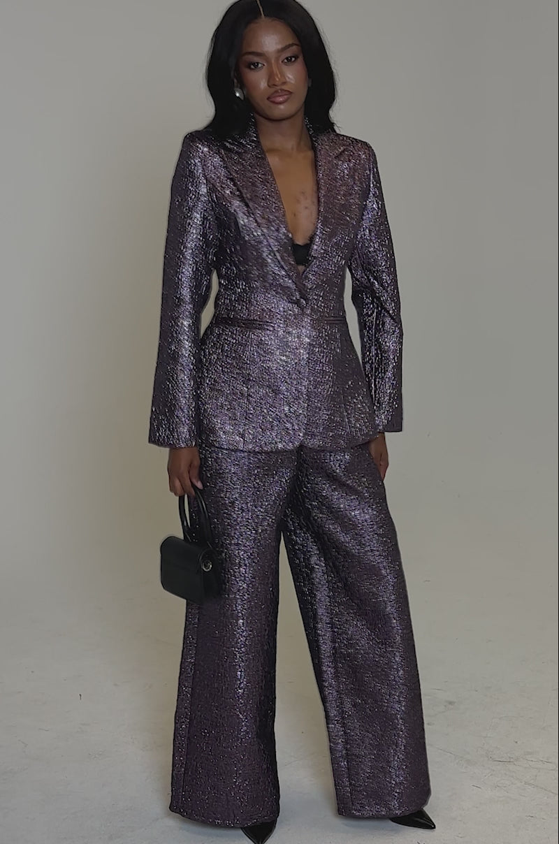 A video of a woman standing confidently against a light background, wearing the HIGH STANDARDS BROCADE BLAZER in textured metallic purple with wide-leg trousers and holding a small black handbag.