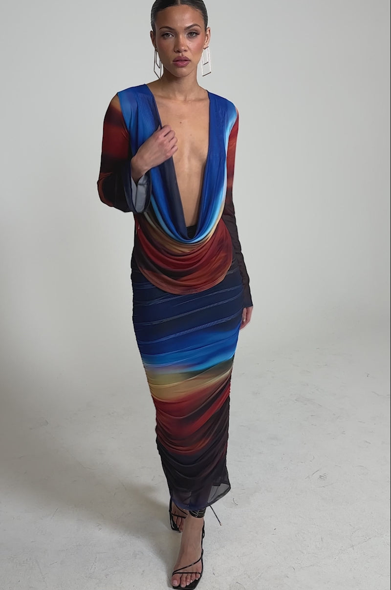 Video of woman modeling the BOOM BOOM MESH MAXI SKIRT, a long ombre maxi skirt with ruched details and a vibrant blue, red, and yellow gradient. She pairs it with black strappy heels and has her hair styled back.