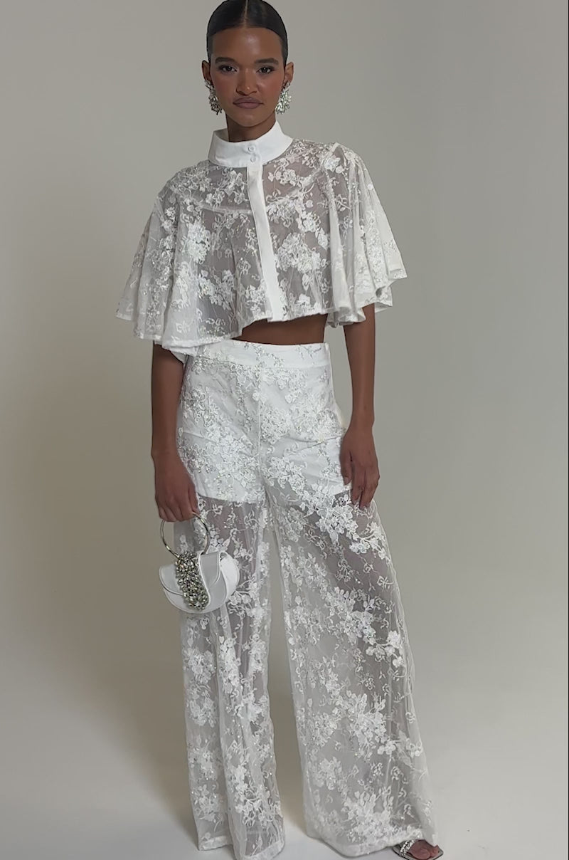 Video of woman modeling the EVA LACE SHORT SLEEVE BUTTON DOWN with high-waisted sheer floral mesh lace trousers, she accessorizes with statement earrings and a small handbag against a plain gray background.
