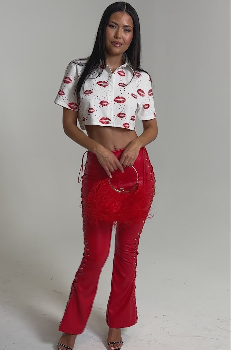 A video of a woman wearing an AKIRA Label KISS KISS RHINESTONE CROPPED BUTTON DOWN, red leather lace-up pants, black high-heeled sandals, and holding a red feathered handbag against a plain light background.