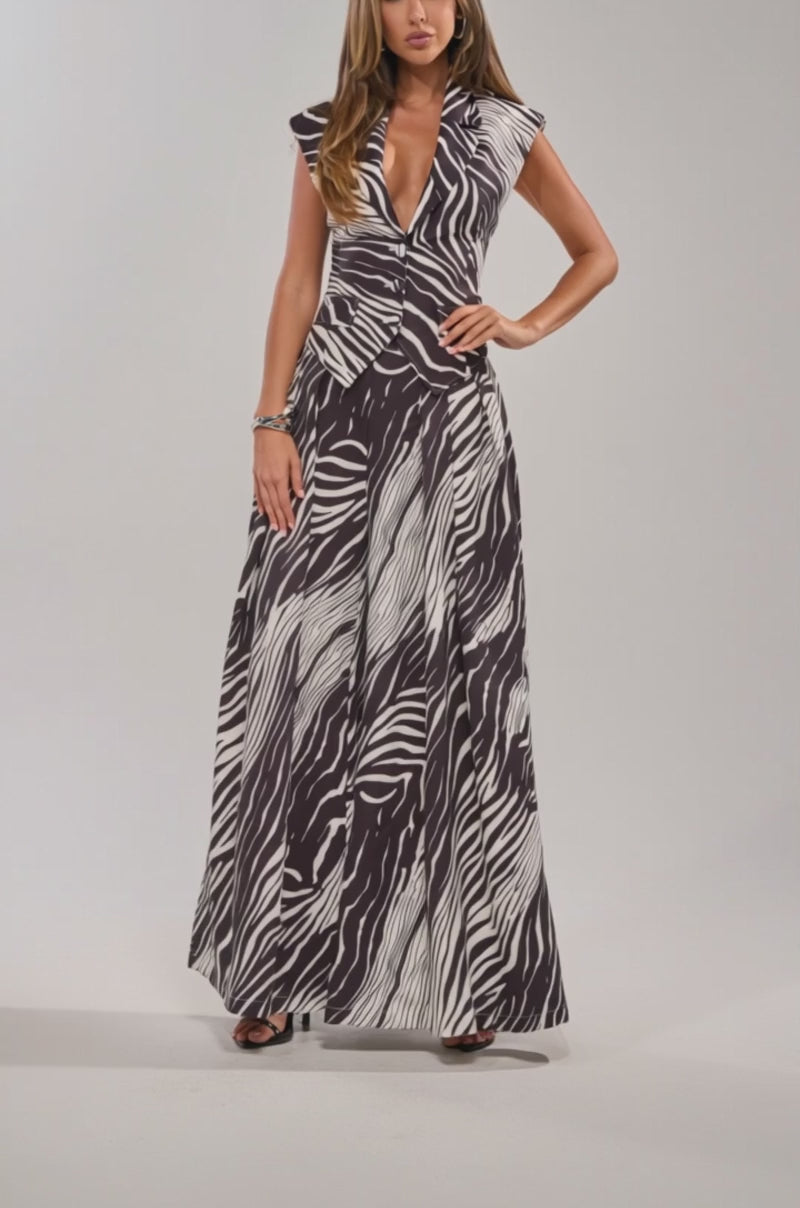 A video of a woman is wearing the WILD AND FREE ZEBRA BUTTON DOWN, a floor-length, tailored zebra-print dress with a deep V-neckline and cap sleeves, standing against a plain light background.