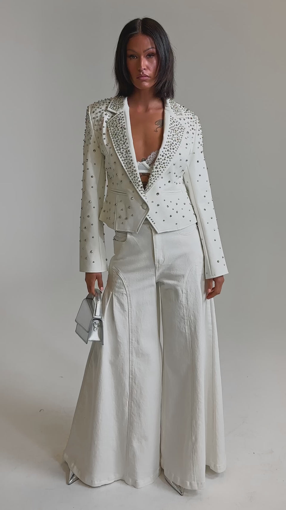 A video of a woman wearing the VIVA VEGAS FITTED MOTO JACKET, wide-leg white pants, a lacy white top, pointed silver shoes, and holding a small metallic silver handbag.