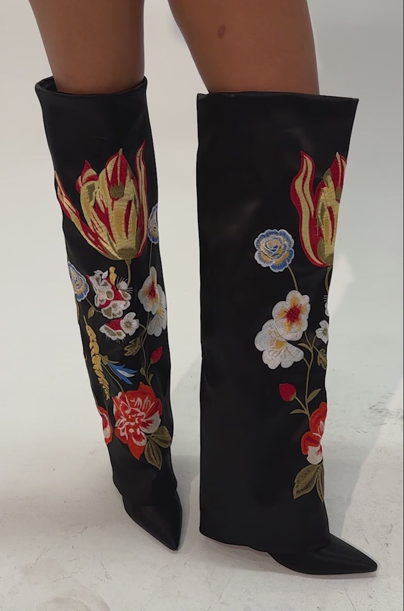 Video of a woman modeling the AZALEA WANG NURIA BLACK MULTI WEDGE EMBROIDERED BOOT—black knee-high boots featuring vibrant floral and leaf embroidery in red, yellow, blue, and white with pointed toes. 