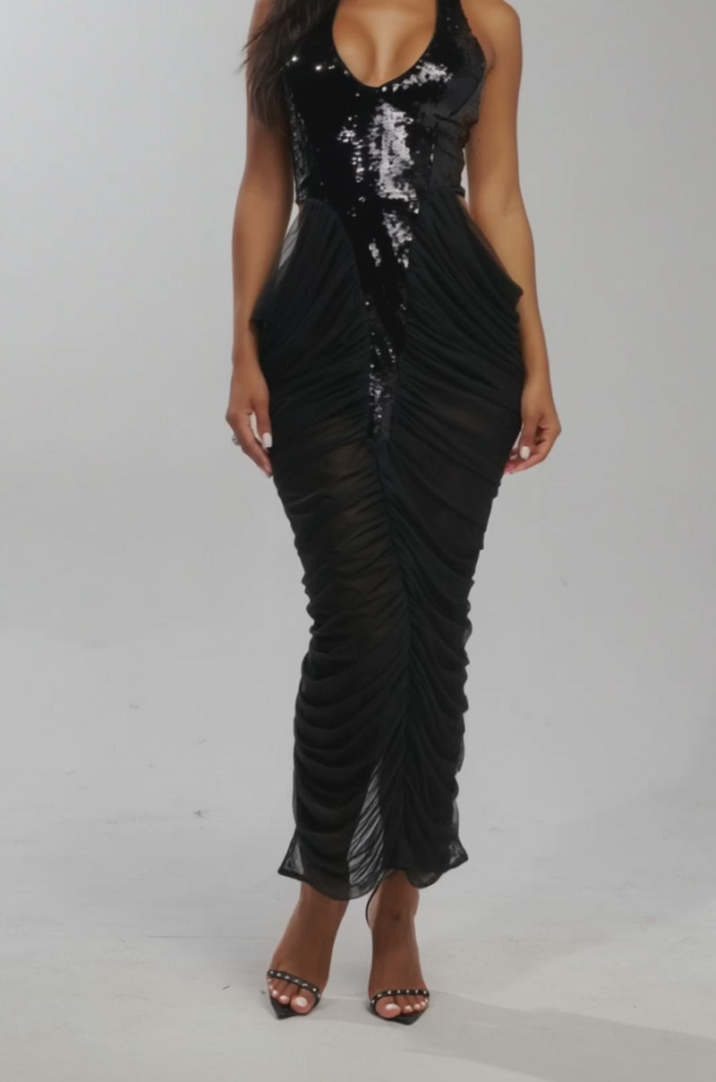 Video of woman modeling the HONEY GLOW SEQUIN MAXI DRESS—a formal black maxi with ruched detailing—paired with open-toe heels against a white background. 