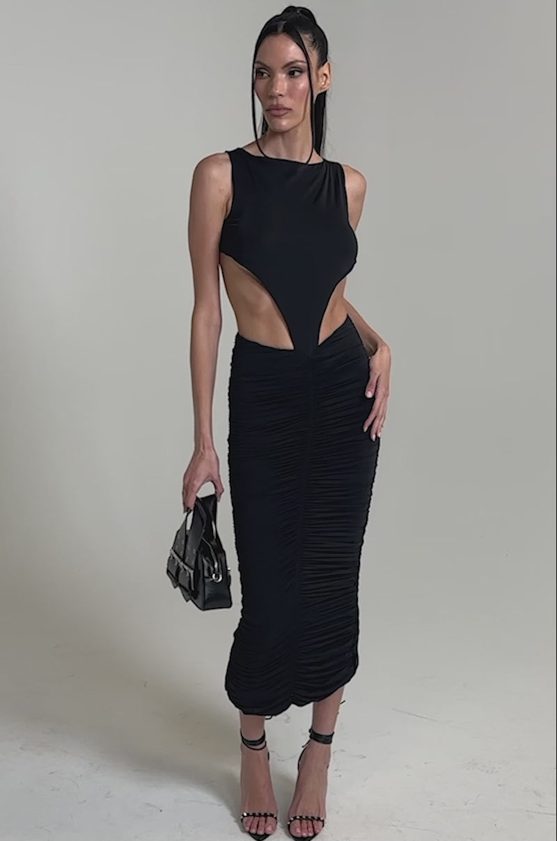 A video of a woman wearing the EYES ON ME MAXI DRESS, a black cutout maxi with a ruched skirt, paired with a small black handbag and strappy heels, standing against a plain background.