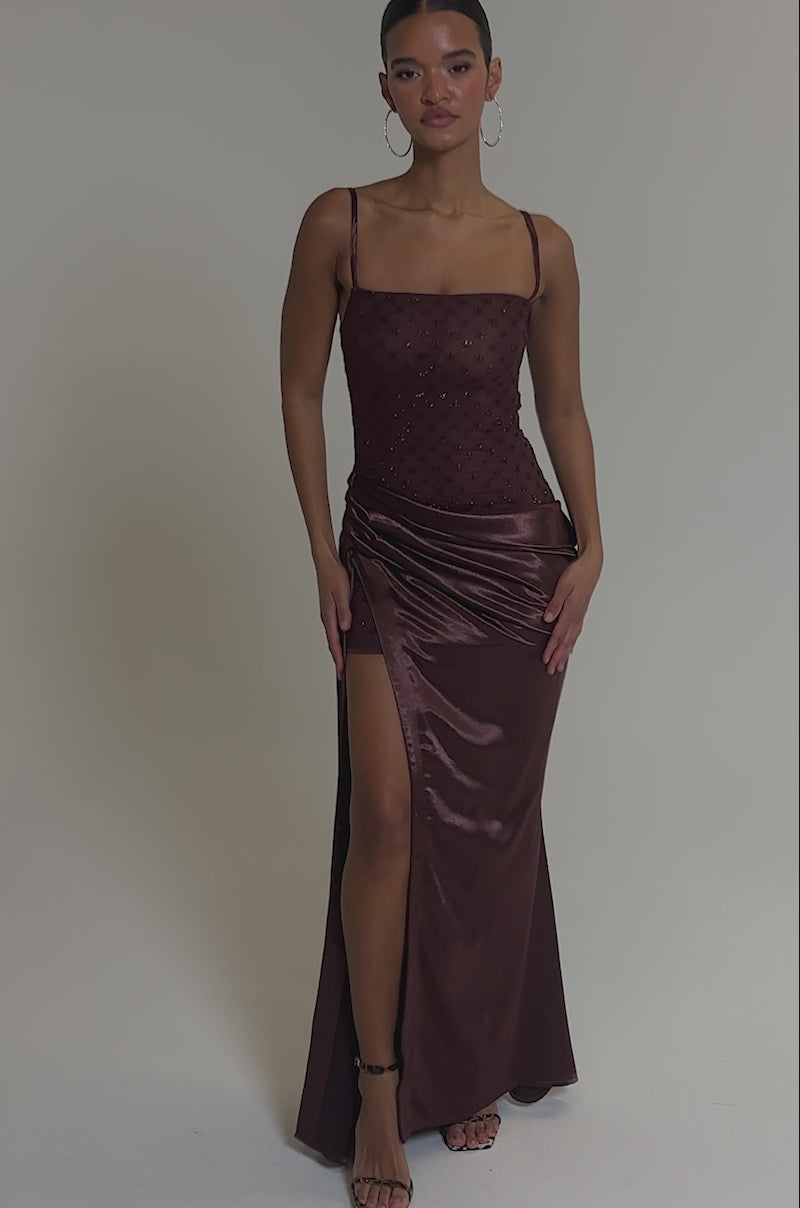 Video of woman modeling the RUNAWAY SATIN MESH MAXI DRESS featuring a patterned bodice, fitted silhouette, high side slit, and ruched details, a woman stands confidently with hoop earrings and ankle-strap heels against a plain background.