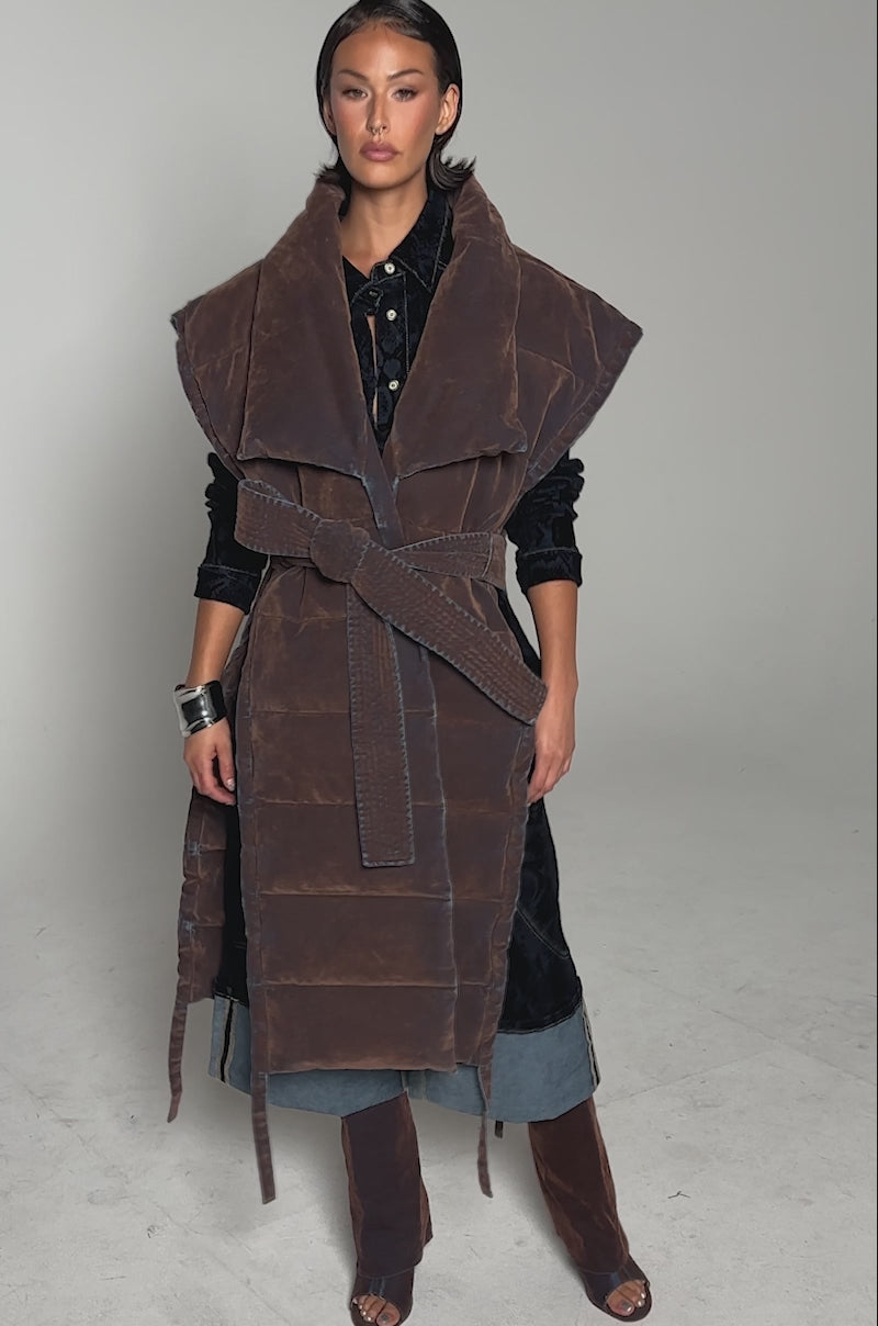 A video of a woman, with short, slicked-back hair, standing against a plain background wearing the PILOT JONES LONGLINE PUFFER VEST IN VELVET DENIM, styled over a dark outfit with denim details and brown open-toe boots.