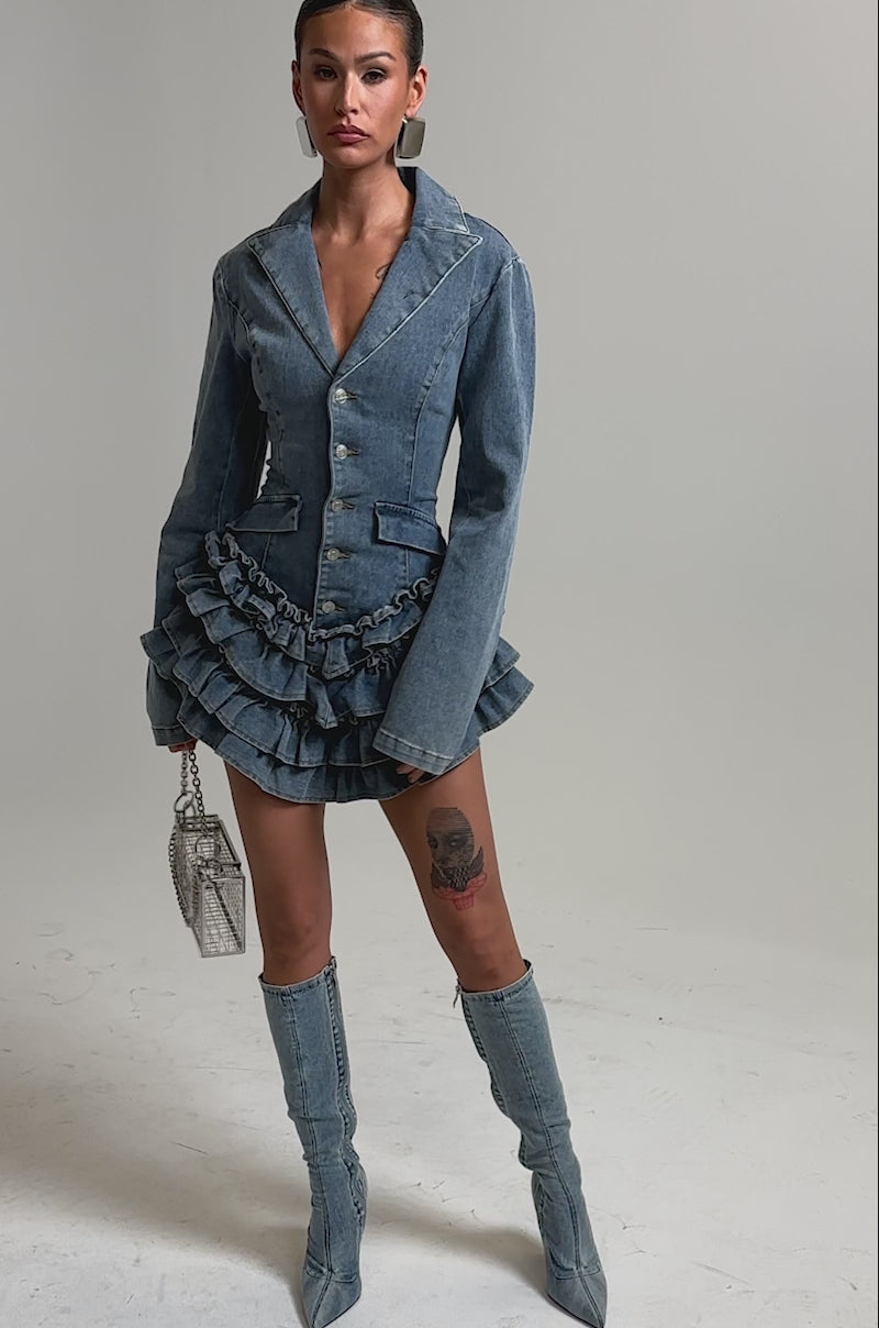 Video of woman modeling the STRICTLY BUSINESS ROMPER IN LIGHT BLUE DENIM with a ruffle mini hem and large silver earrings, standing against a plain background while holding a chain-strap purse.