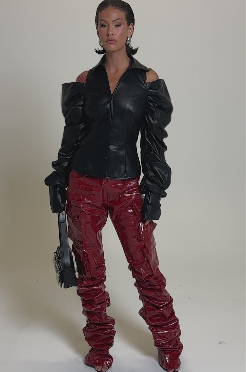 A video of a woman in the CRAZY IN LOVE BLACK FAUX LEATHER BUTTON DOWN BLOUSE with puff sleeves, paired with shiny red ruched vinyl pants, a black handbag, and heeled open-toe shoes against a plain light gray background.