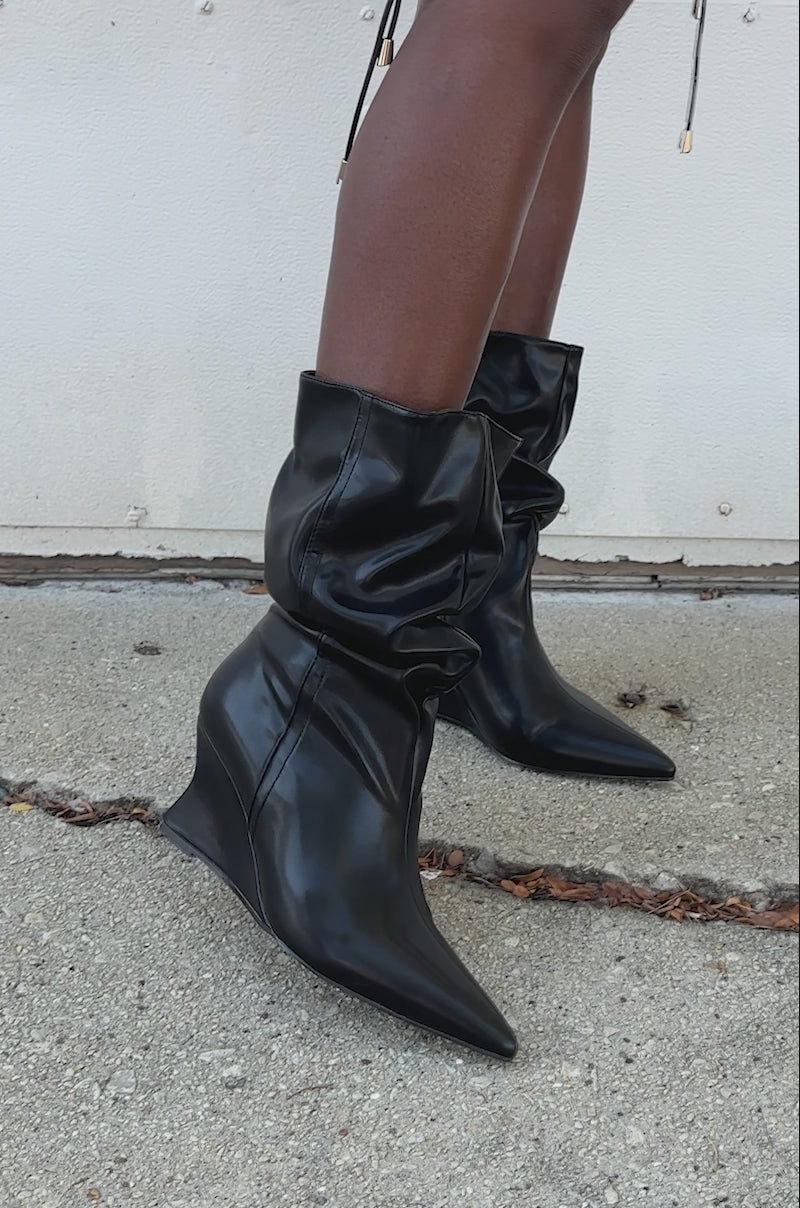 A video of a person wearing AZALEA WANG ASAF Ruched Wedge Bootie in Black with a slouchy, shiny design, standing on concrete with dried leaves nearby.