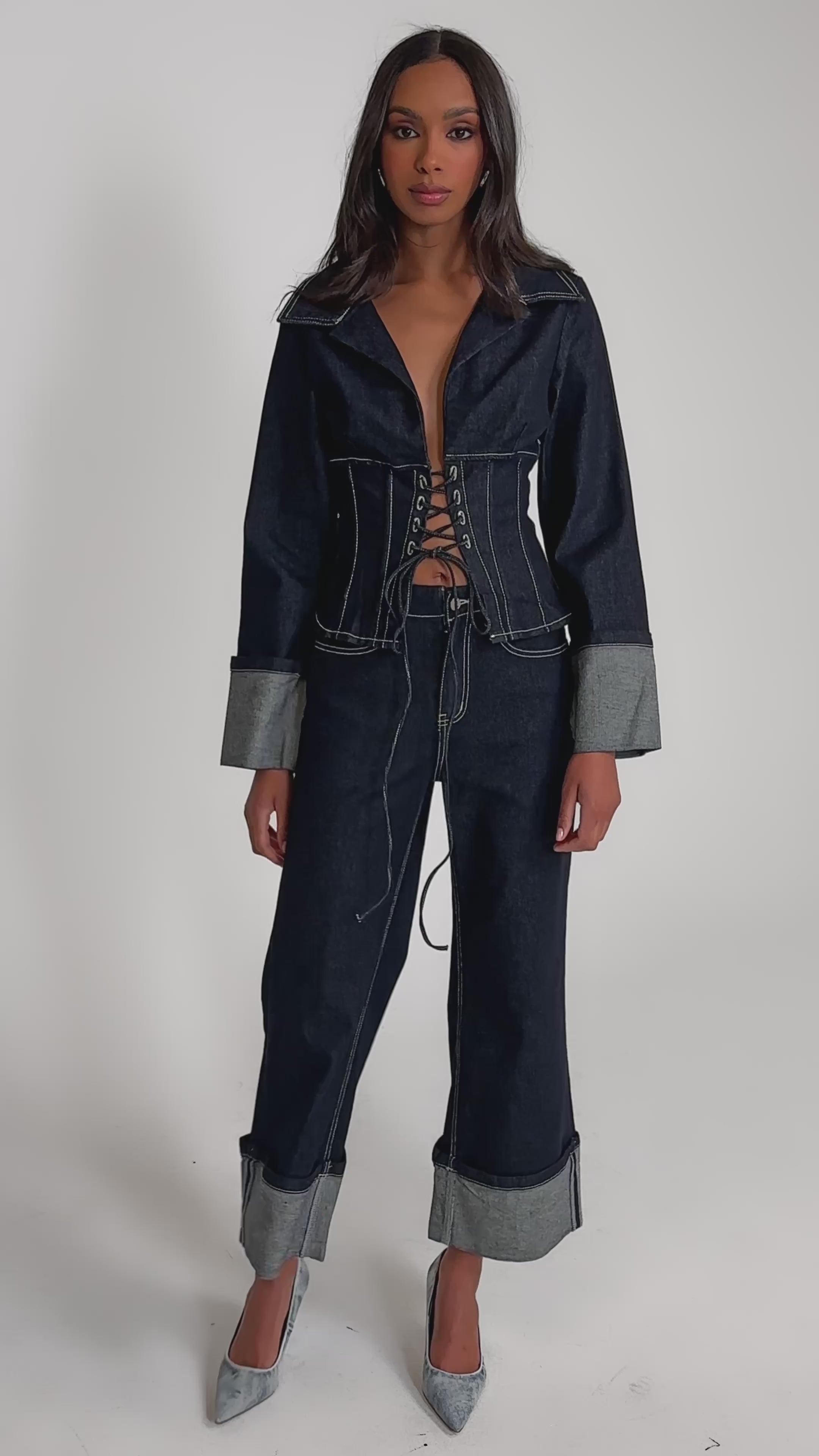 Video of woman modeling the DON'T TALK TO ME LACE UP DENIM BLOUSE, featuring tailored wide cuffed sleeves and a corset-style lace-up front, standing against a plain background with a serious expression.